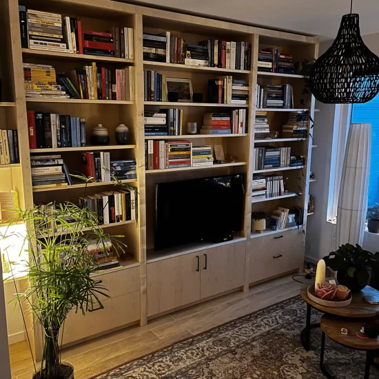 Een lichtbruine houten boekenkast gemonteerd in een woonkamer. De boekenkast bestaat uit drie delen. Het linker en rechtergedeelte van de boekenkast heeft onderin twee lades met daarboven boekenplanken die vol liggen met boeken. Het midden gedeelte heeft onderin twee deurtjes, daarboven staat een televisie en boven de televisie vier boekenplanken.