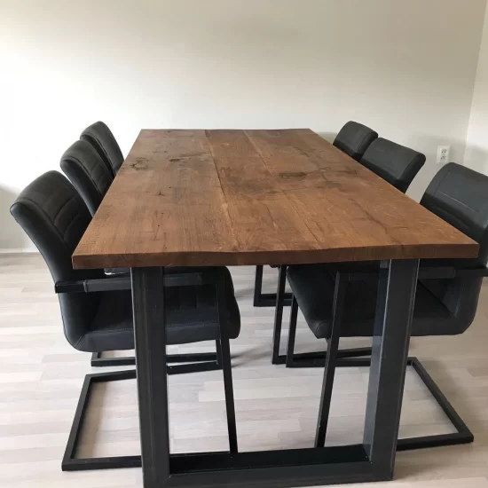 Een lang rechthoekige eettafel met een donkerbruin grof houten blad met een zwart stalen frame. Aan beide zijden van de tafel staan drie zwarte beklede eetstoelen.