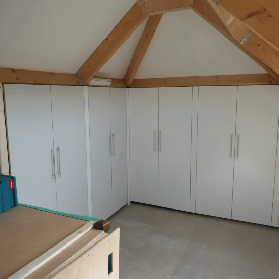 Vier grote witte kledingkasten met klapdeuren en grote chroomkleurige handvatten. De kledingkasten zijn verwerkt in schuin aflopende wanden van een zolderruimte.