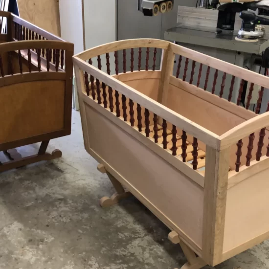 Twee houten ledikantjes met bovenin een doorkijkrand met kleine houten spiralen. De ledikantjes hebben de vorm van een omgekeerde trapeze, zijn daarom smaller aan de onderkant en staan op een houten frame.