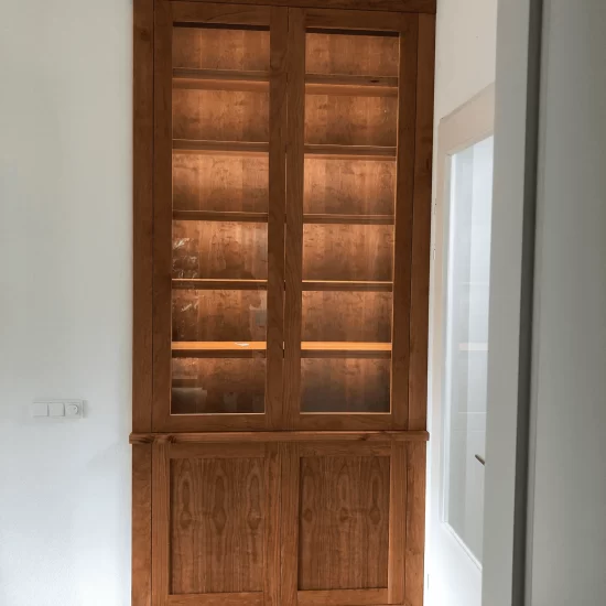 Een donkerbruine vitrinekast, ingebouwd in een nis. De kast maakt gebruik van magneetsluitingen, heeft onderin twee houten deurtjes en daarboven twee lange deuren met doorzichtig glas. Achter het glas zijn vijf houten planken zichtbaar die worden verlicht.